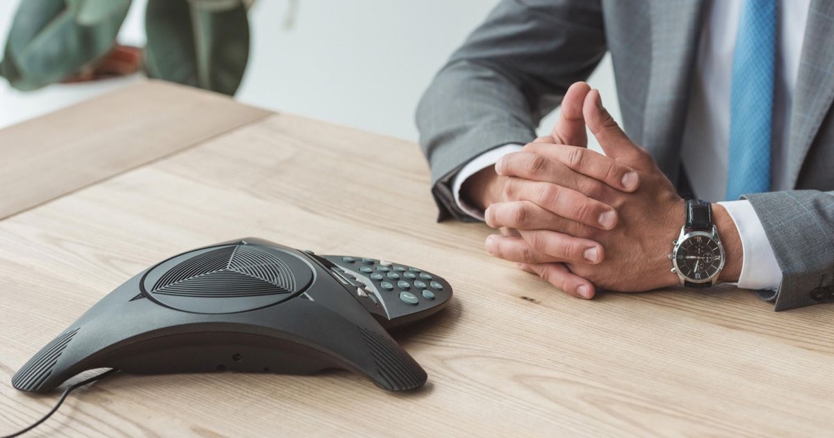 Ein Konferenztelefon steht auf einem Tisch. Daneben sitzt ein Geschäftsmann mit gefalteten Händen, der an einer Telefonkonferenz teilnimmt.