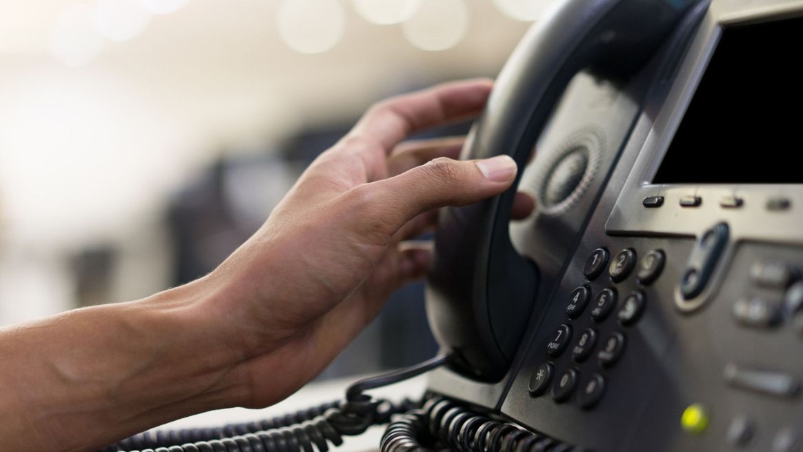 Ausschnitt eines modernen Festnetztelefons in einem Büro. Eine weibliche Hand bedient eine Taste. Davon ausgehend erscheinen im Vordergrund virtuelle Symbole für Telefonkonferenzen und andere Funktionen.