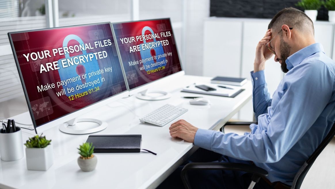 Ein resignierter Mann sitzt vor zwei Monitoren, auf denen Symbole von Schlössern eine durch einen Trojaner verursachte kriminelle Verschlüsselung der Daten darstellen.
