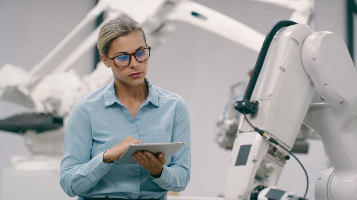 Eine Frau steht mit einem Tablet in der Hand neben einem Roboterarm und schaut sich diesen an
