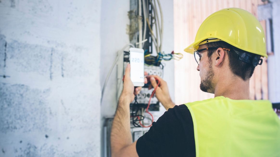 Ein Mann mit einem Helm kontrolliert auf einer Baustelle einen Verteilerkasten. In der linken Hand hält er ein Smartphone, das Messwerte anzeigt.