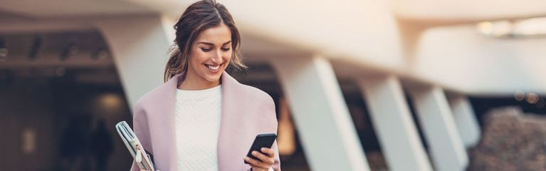 Eine Frau mit einer rosa Jacke steht im Freien vor einem Gebäude und betrachtet lächelnd ihr Mobiltelefon