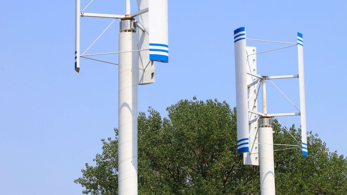 Zwei Windkraftanlagen mit Darrieus-H-Rotor vor einem Baum und blauem Himmel
