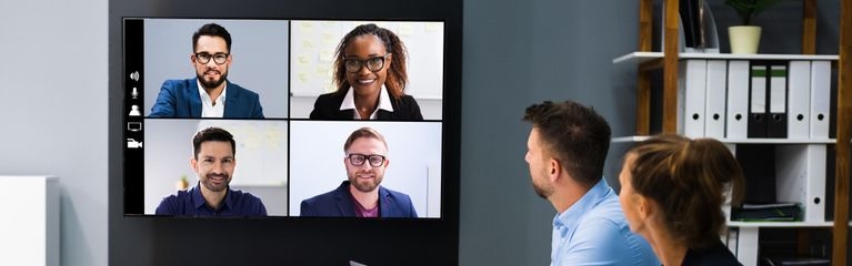 Zwei Männer und zwei Frauen sitzen in einem Besprechungszimmer und nehmen an einer Videokonferenz teil. Auf einem Bildschirm betrachten sie vier andere Teilnehmer:innen der Videokonferenz.