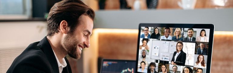 Ein Mann sitzt lächelnd vor einem Monitor, auf dem die Teilnehmer:innen einer Videokonferenz zu sehen sind