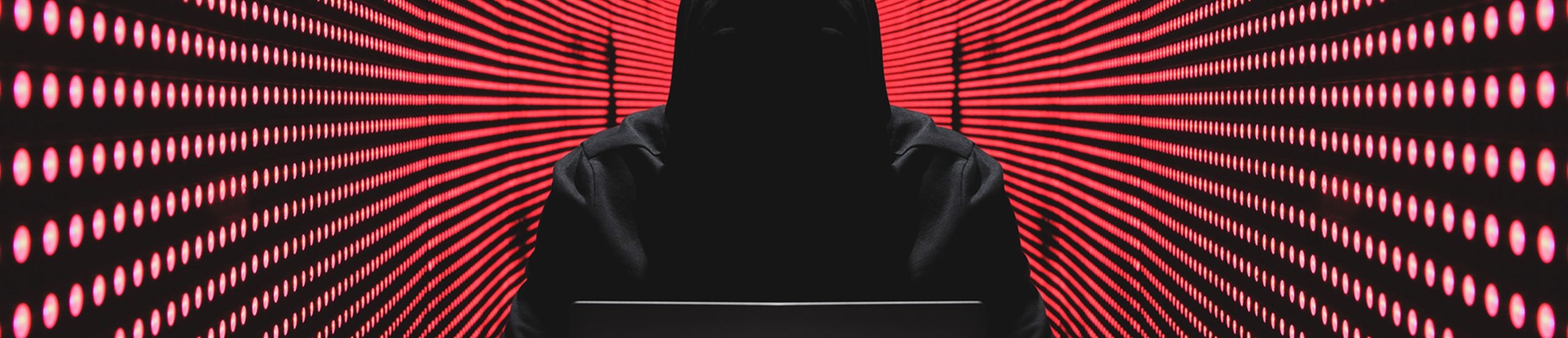 Person mit schwarzem Kapuzenpullover sitzt vor einem Laptop. Darum sind rote runde Lichter an einer schwarzen Wand