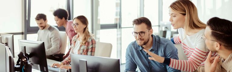 Drei Männer und eine Frau sitzen an einem Tisch vor Computermonitoren. Dahinter stehen zwei Frauen, eine deutet auf einen Bildschirm