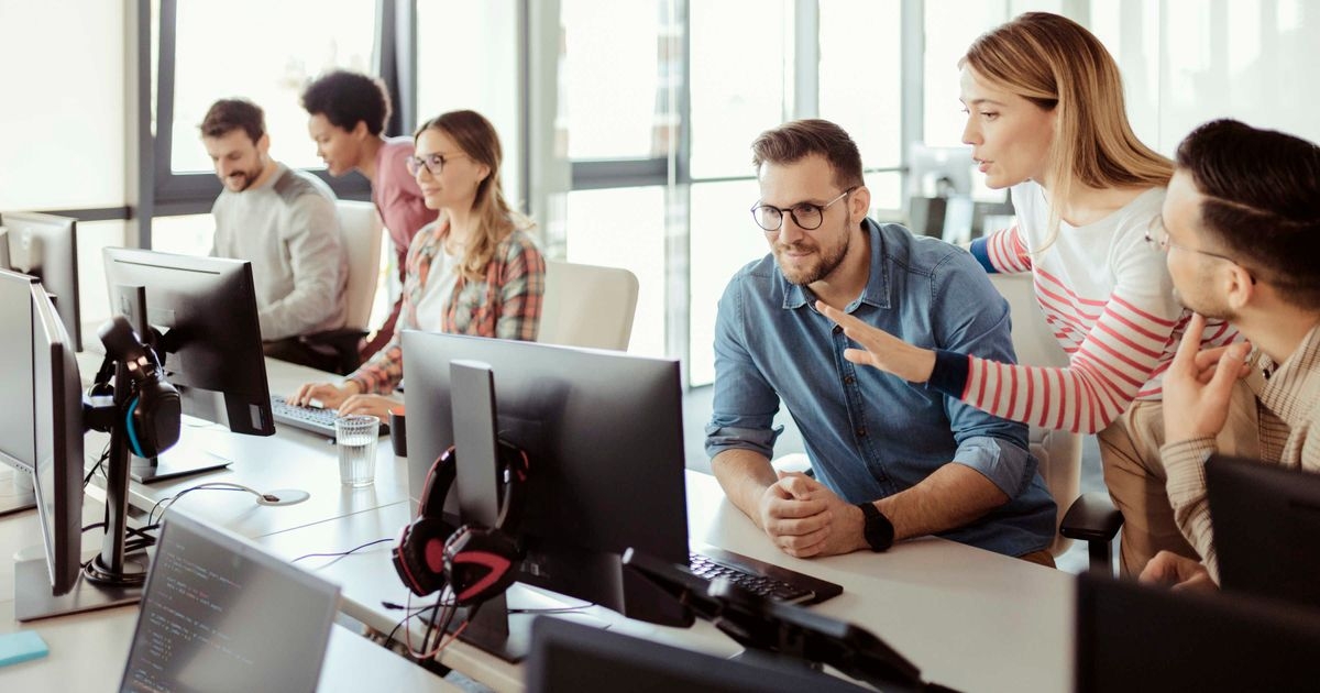 Drei Männer und eine Frau sitzen an einem Tisch vor Computermonitoren. Dahinter stehen zwei Frauen, eine deutet auf einen Bildschirm