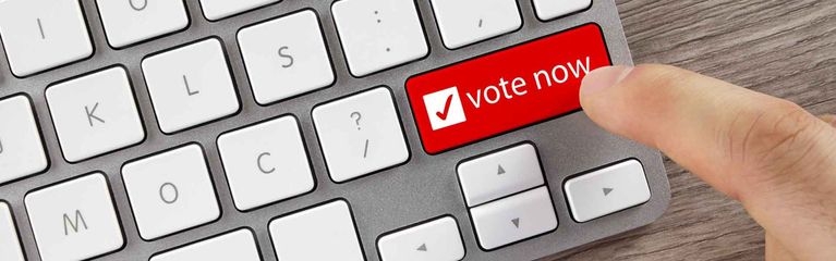 Ein Finger bewegt sich auf eine mit “vote now” beschriftete Taste auf einem Notebook zu.