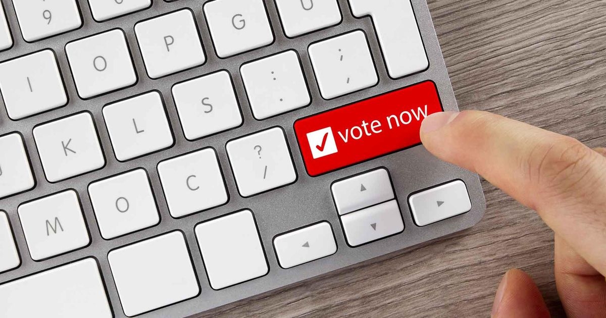 Ein Finger bewegt sich auf eine mit “vote now” beschriftete Taste auf einem Notebook zu.
