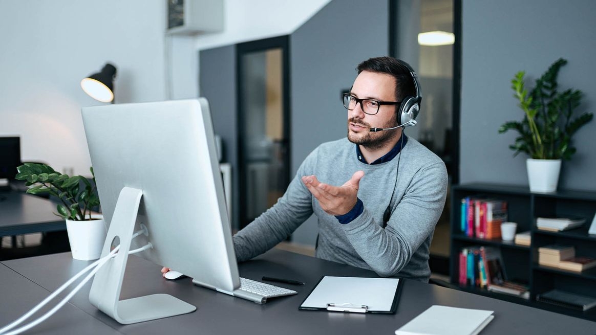 Ein Mann sitzt in einem Büro und telefoniert mit einem Headset. Dabei blickt er auf einen Monitor auf dem Schreibtisch.