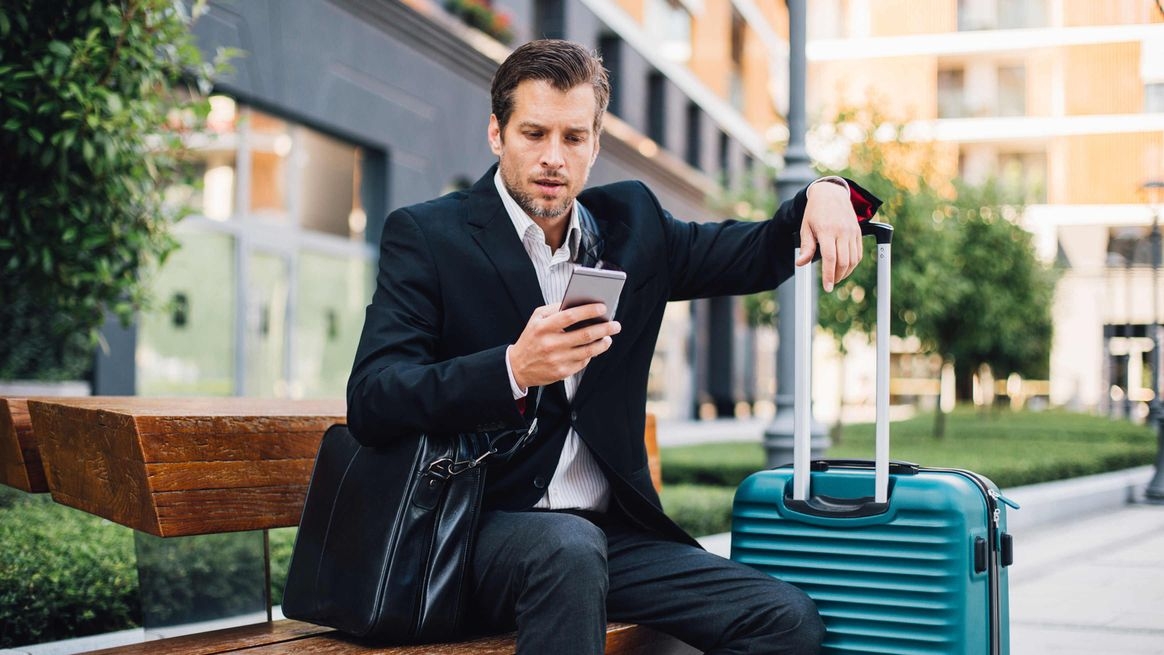 Ein Mann mit Gepäck sitzt draußen auf einer Bank und schaut auf sein Smartphone