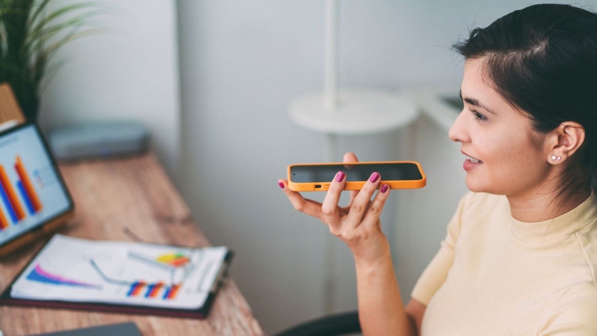 Eine Frau sitzt an ihrem Arbeitsplatz im Büro und spricht eine Sprachnachricht auf ihrem Smartphone auf.