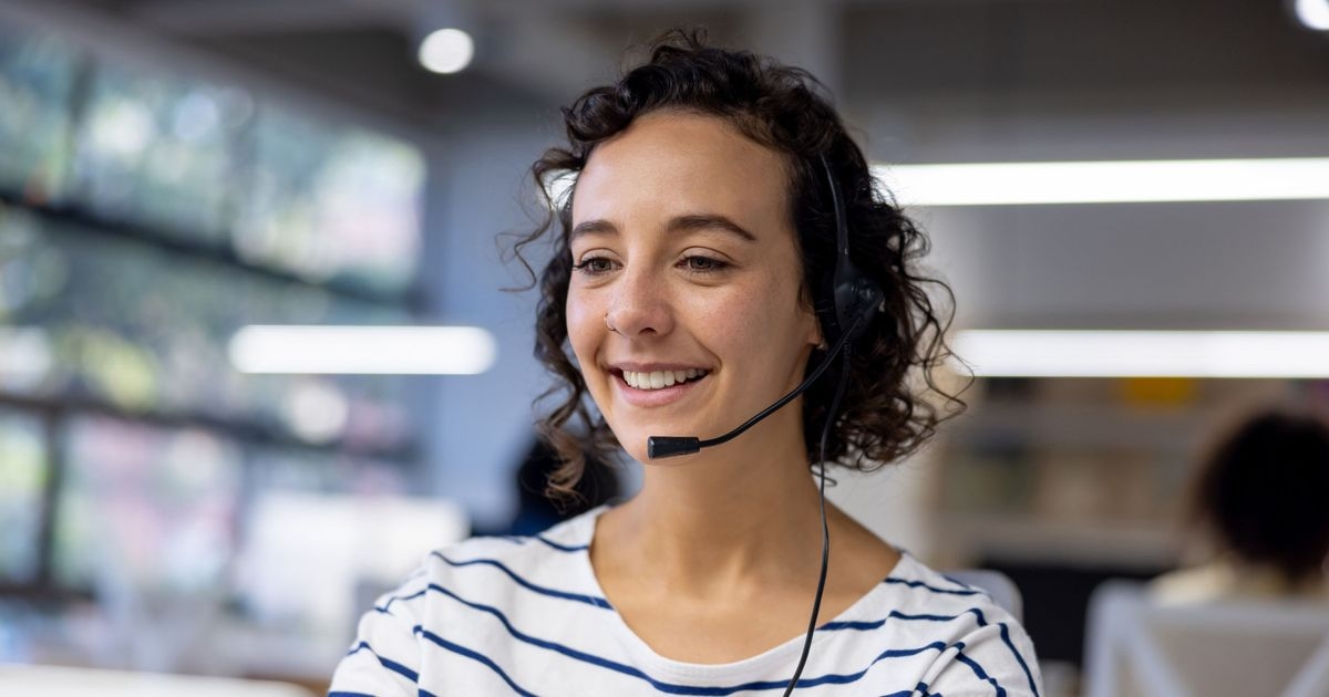 Eine junge Frau trägt ein Headset und schaut lächelnd auf einen Monitor.