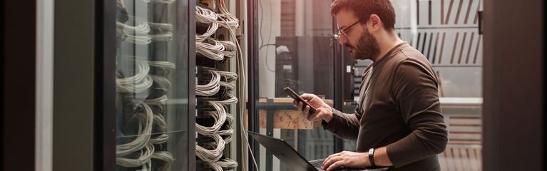 Mann kniet mit Laptop und Handy vor einem Server-Schrank, aus dem viele Kabel herausragen