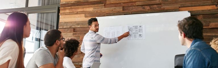 Mann zeigt auf Zettel an einem Whiteboard, davor sitzen mehrere Zuhörer:innen an einem Konferenztisch.