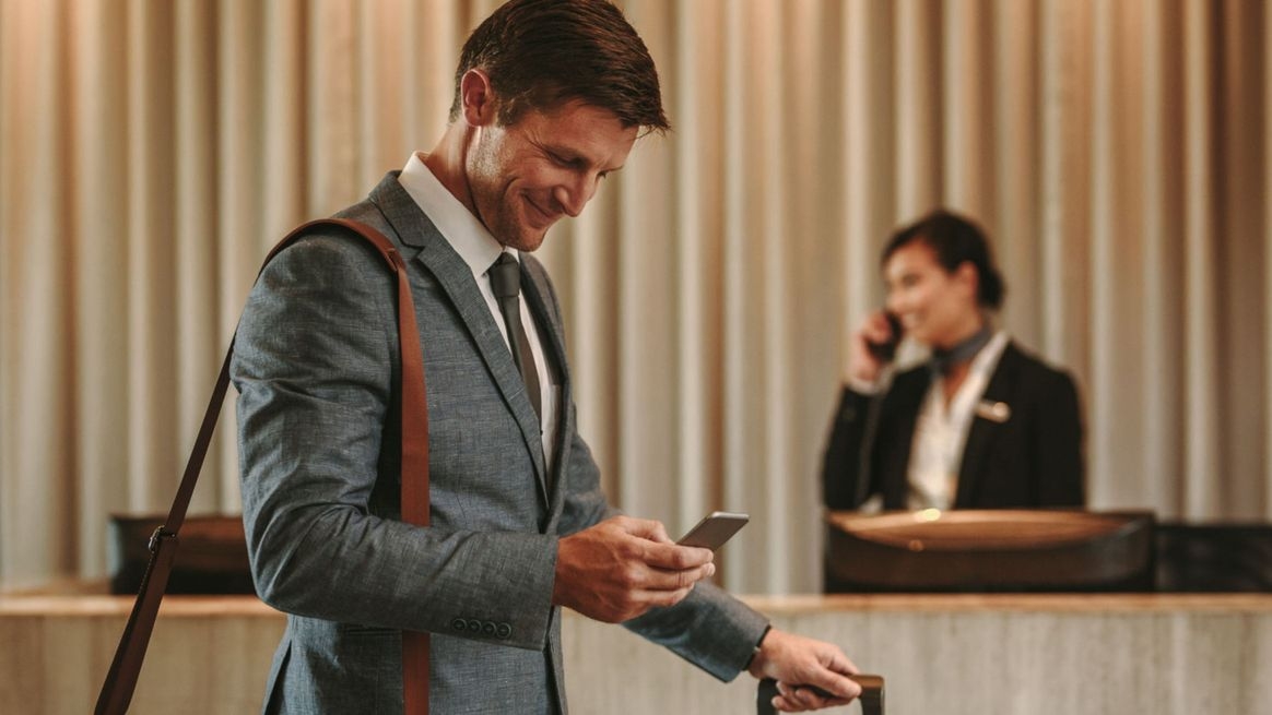 Ein Mann Mann im Anzug mit Rollkoffer und Tasche über der Schulter guckt lächelnd auf sein Handy, im Hintergrund telefoniert eine Frau des Bodenpersonals