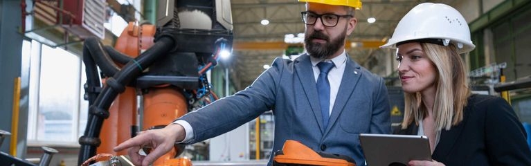 Ein Mann und eine Frau inspizieren in einer Industriehalle eine Maschine. Der Mann zeigt auf etwas. Die Frau hält einen Laptop in den Händen, der Mann in der linken Hand ein Diagnosegerät.