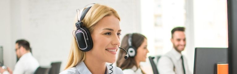 Eine lächelnde Frau spricht in ein Headset und schaut auf einen Computerbildschirm.