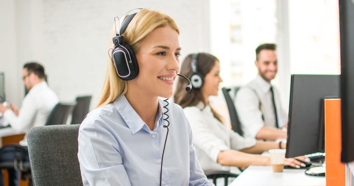 Eine lächelnde Frau spricht in ein Headset und schaut auf einen Computerbildschirm.