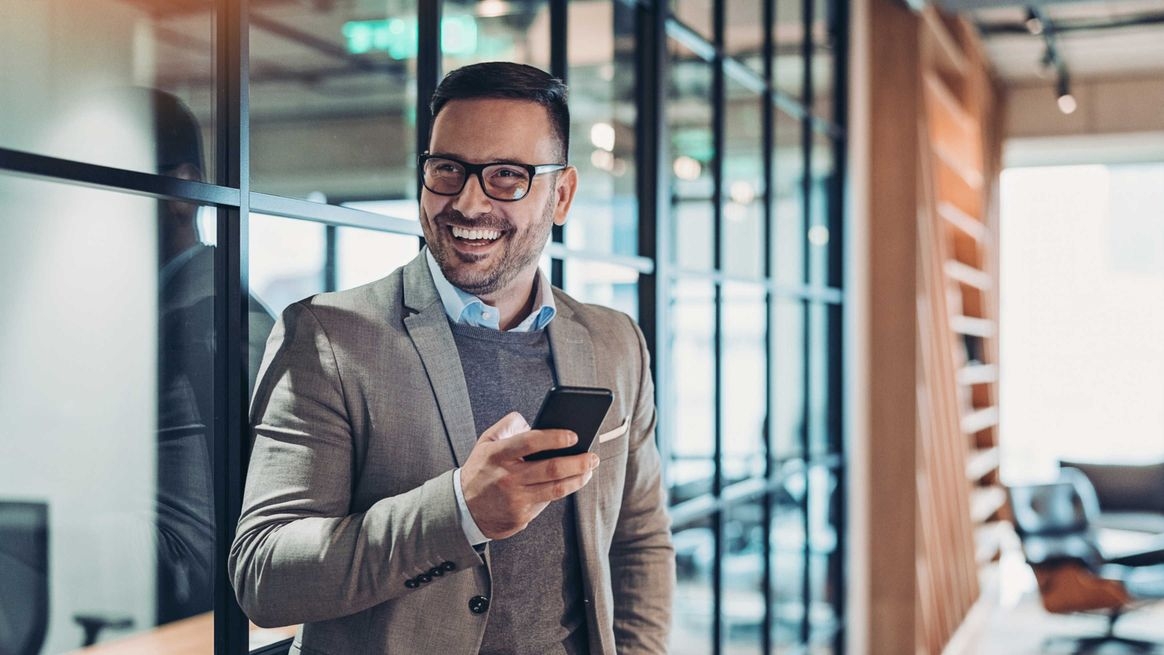 Person mit Smartphone in der Hand steht in einem Büro und lacht in die Kamera