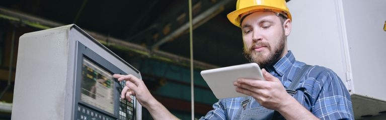 Ein Mann mit gelbem Schutzhelm bedient mit einer Hand eine Fabrikmaschine und hält in der anderen Hand ein Tablet.