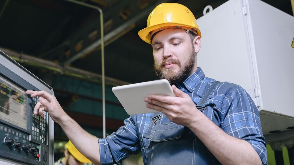 Ein Mann mit gelbem Schutzhelm bedient mit einer Hand eine Fabrikmaschine und hält in der anderen Hand ein Tablet.