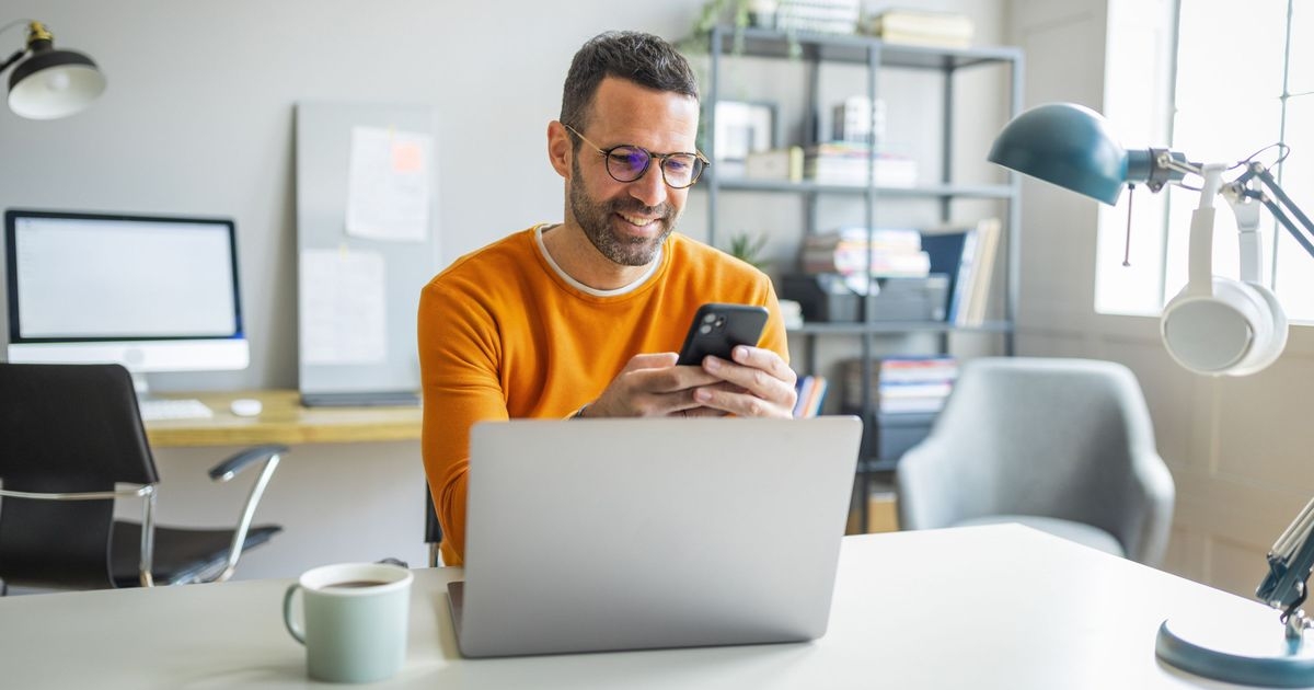 Ein Mann sitzt an einem Schreibtisch und blickt lächelnd auf ein Smartphone. Vor ihm stehen ein Notebook und eine Tasse.