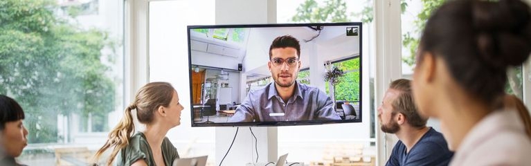 Drei Frauen und ein Mann sitzen an einem Tisch und blicken zu einem Monitor, auf dem ein Mann zu sehen ist. Auf dem Tisch stehen Notebooks.