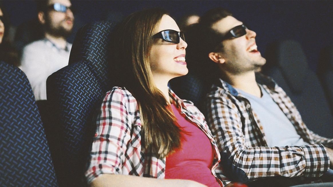 Ein Mann und eine Frau mit 3D-Brillen sitzen in Sesseln. Hinter ihnen sitzen weitere Personen