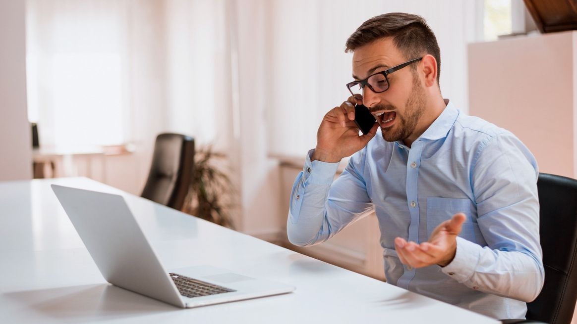 Mann in Businesskleidung sitzt telefonierend vor einem Notebook und gestikuliert dabei.