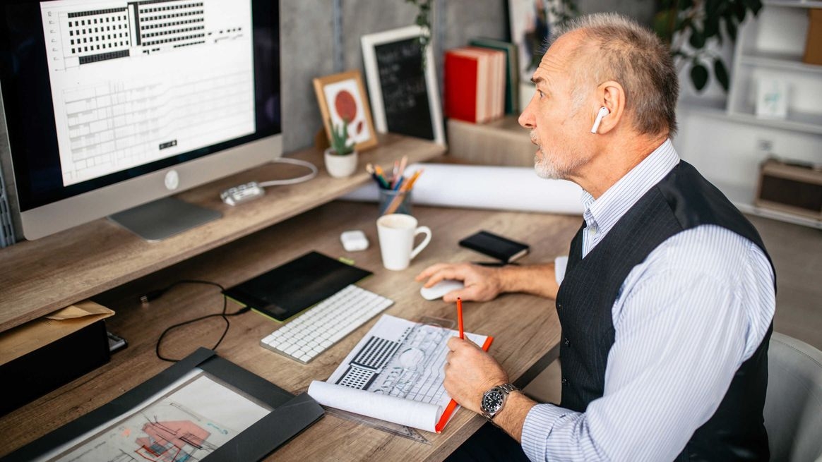 Ein älterer Mann sitzt an einem Schreibtisch vor einem Monitor. Auf dem Bildschirm und auf dem Tisch sind Grafiken und Skizzen zu sehen.  