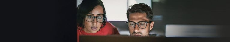 Zwei Personen sitzen vor Computermonitoren in einer Büroumgebung und betrachten konzentriert Bildschirminhalte. Die Monitore beleuchten die Gesichter, während der Hintergrund unscharf dargestellt ist.