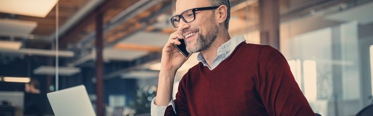 Mann sitzt im Büro am Laptop und telefoniert mit dem Smartphone.