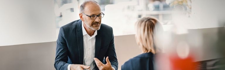 Das Bild zeigt zwei Personen in Businesskleidung, die in einem hellen, modern aussehenden Raum miteinander sprechen. Die Szene wirkt wie ein Gespräch in einem professionellen oder geschäftlichen Umfeld.