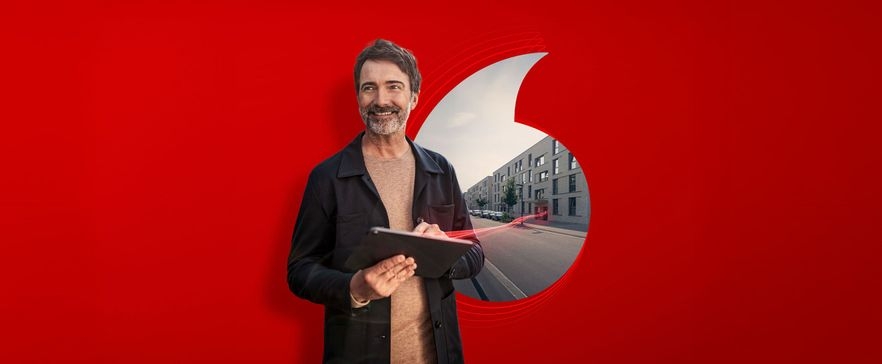Person steht vor rotem Hintergrund mit Vodafone‑Logo und hält ein Tablet vor einem urbanen Straßenmotiv.
