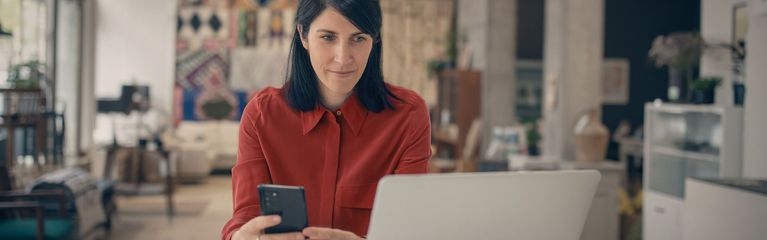 Frau sitzt mit Smartphone am Laptop.