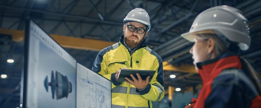 Ein Bauingenieur steht mit seinem Tablet neben einer Kollegin am Computer